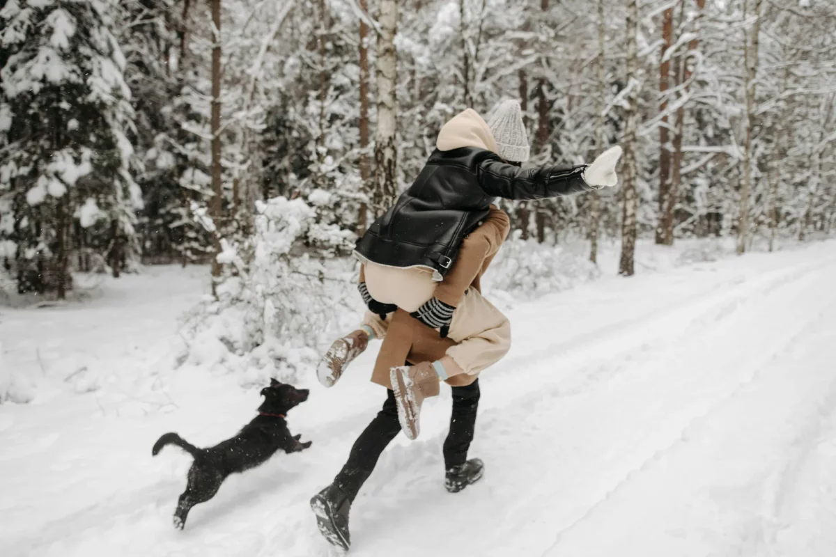 Ein Paar genießt den Winter im Schnee mit einem Hund – Vitamin D Mangel vorbeugen durch frische Luft und Bewegung.
